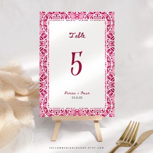 May include: A wedding table number card on a small wooden easel. The card has a white background with a decorative magenta border. The number "5" is prominently displayed, along with the names "Parisa + Omar" and the date "23.11.2X".
