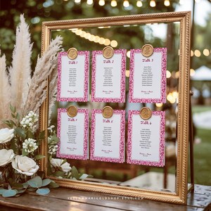 May include: A gold framed seating chart with six cards, each with a pink floral border and gold wax seals. The cards display guest names and table numbers. The background includes flowers and greenery.