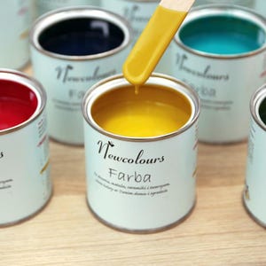 Könnte beinhalten: Eine Auswahl kleiner Farbtöpfe in verschiedenen Farben, darunter Rot, Gelb, Blau und Grün. Die Dosen sind mit "Newcolours Farba" beschriftet und werden mit einem Holzstab getestet. Die Farbtöpfe sind weiß mit einem hellblauen Etikett.