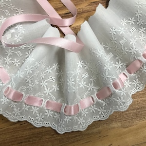 Könnte beinhalten: Weiße Baumwollspitzenborte mit einem gewellten Rand und rosa Satinband-Bändern. Die Spitze hat ein zartes Blumenmuster.