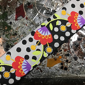 Puede incluir: Tira de tela decorativa con un estampado floral. El diseño incluye flores bordadas rojas, moradas y amarillas sobre un fondo de lunares blancos y negros. Hojas verdes y detalles amarillos completan el diseño.