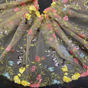 May include: Delicate floral embroidered fabric in shades of yellow, pink, purple, and blue. The embroidery features a variety of flowers and leaves on a sheer, beige background. This fabric is suitable for apparel and craft projects.