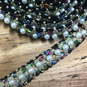 May include: Black and gold trim with white pearl beads. The trim has a woven texture and is decorated with colorful threads.