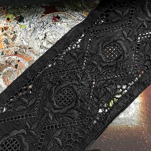 May include: Black embroidered trim with a floral and geometric pattern. The trim has cut-out details and small, reflective embellishments. The fabric looks like a soft, woven material, suitable for craft projects.