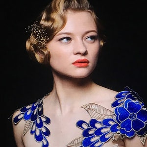 May include: A woman with a vintage hairstyle wears a blue and gold embroidered neckline with a large blue flower on one shoulder.