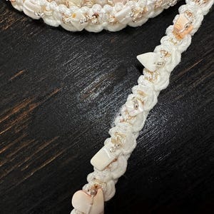 May include: Close-up of a white and cream-colored braided headband with decorative shells and silver beads. The headband is on a dark wooden surface. The design features a repeating pattern of shells and beads.
