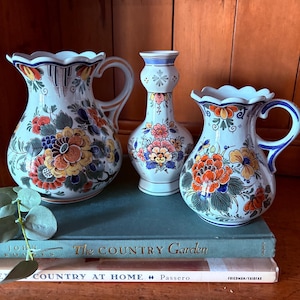 May include: Three ceramic vases with floral designs. The vases are white with blue, orange, yellow, and green floral patterns. The vases are sitting on top of books with the titles "The Country Garden" and "Country at Home".