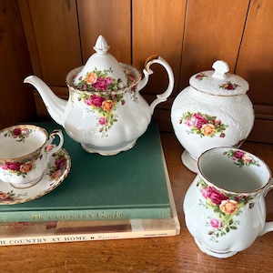 May include: A white porcelain tea set with a floral rose pattern. The set includes a teapot, sugar bowl, teacup, saucer, and mug. The tea set is decorated with pink, yellow, and orange roses and gold trim.