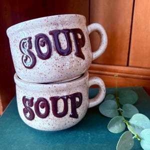 May include: Two stacked soup mugs with a speckled, off-white finish. The word "SOUP" is prominently displayed in a dark brown, stylized font on each mug. Each mug has a handle.