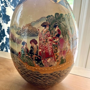 May include: A large, egg-shaped decorative ceramic vase with a detailed painted scene of figures in traditional clothing. The vase features a cream-colored background with gold accents and a depiction of a boat with people. The base is a dark brown color.