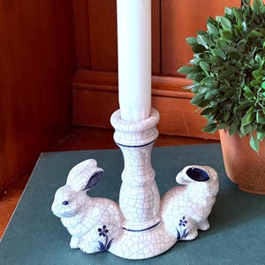 May include: A white ceramic candle holder with a crackle glaze design, featuring two bunny figures on either side. The bunnies have blue accents on their ears and a small blue flower detail. A white candle is inserted in the center.