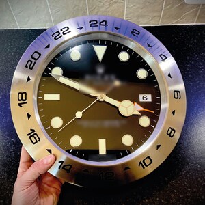 May include: A silver wall clock with a black face and white numbers. The clock has a blue bezel with white markings and a silver outer ring with black numbers.