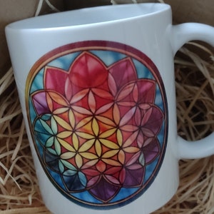 Könnte beinhalten: Eine weiße Keramiktasse mit einem farbenfrohen geometrischen Design des Lebensblume-Symbols. Die Tasse hat einen Henkel und steht auf einer braunen Oberfläche.