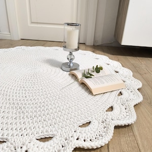 Op de afbeelding: Een rond, wit gehaakt kleed met een gestructureerd, dicht steekpatroon in het midden en een decoratieve geschulpte rand met opengewerkte details. Het kleed is gemaakt van dik, zacht garen en ligt plat, wat zorgt voor een neutrale uitstraling.
