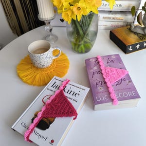 Puede incluir: Una imagen de naturaleza muerta con dos libros con marcapáginas en forma de bikini de ganchillo. Uno es rosa oscuro, el otro rosa claro. También se ven un jarrón con narcisos amarillos, una taza de café y un posavasos de macramé amarillo.