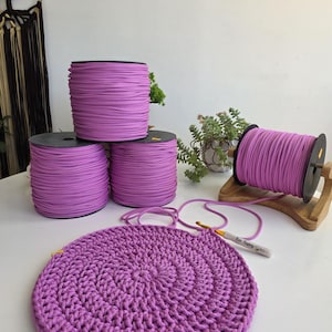 Può includere: Quattro bobine di corda color lavanda sono disposte con tre impilate e una su un supporto in legno. Un pezzo circolare all'uncinetto color lavanda si trova in primo piano con un uncinetto argento e oro appoggiato sopra, con il testo Bee Happy.