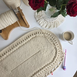 Puede incluir: Una alfombrilla de ganchillo de color crema con forma ovalada y borde decorativo, un ganchillo rosa con botones de colores, bobinas de cordón crema en un soporte de madera, un jarrón de cristal con dos rosas rojas y una taza blanca con café.