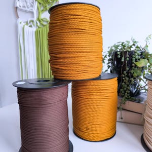 Puede incluir: Tres carretes de cordón, uno marrón, uno amarillo mostaza y uno beige. Los carretes están sobre una superficie blanca.