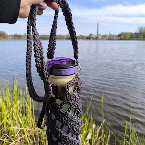 May include: A crocheted water bottle holder in purple and grey tones. The holder has a long strap for carrying over the shoulder.