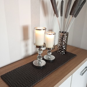Könnte beinhalten: Zwei silberne Kerzenhalter mit klaren Glaskerzenbehältern und weißen Kerzen. Die Kerzenhalter stehen auf einem dunkelgrauen gewebten Tischläufer auf einer hellbraunen Oberfläche. Eine Vase mit grauen getrockneten Schilf steht im Hintergrund.