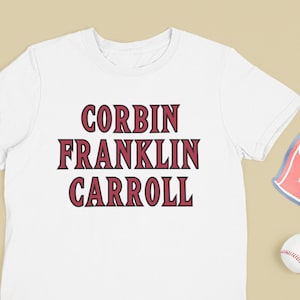 May include: White t-shirt with the text "CORBIN FRANKLIN CARROLL" in maroon. A baseball bat, a baseball glove, a baseball, a foam finger with the text "WE'RE #1", and a turquoise baseball cap are arranged around the t-shirt.
