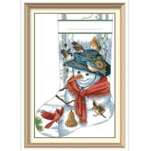 May include: Cross-stitch artwork of a snowman inside a Christmas stocking. The snowman wears a blue hat, red scarf, and has a carrot nose. Birds perch on the hat and around the stocking. A bell and a red bird are also featured.