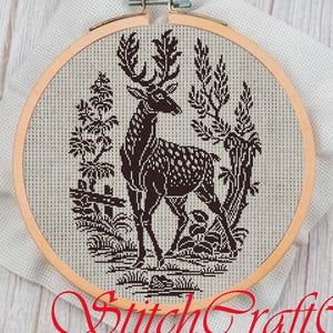 May include: Black and white cross-stitch embroidery of a deer standing in a forest setting. The deer is surrounded by trees and plants. The embroidery is framed in a wooden hoop.
