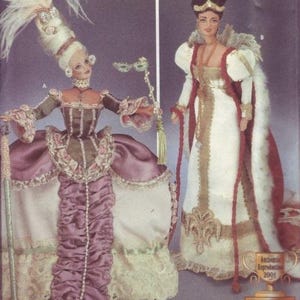 May include: Two dolls in historical costumes. One doll wears a tall white wig, a brown and pink dress with lace trim, and holds a mask and staff. The other doll is in a white and red gown with gold accents and a crown.