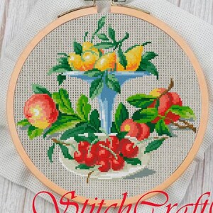 Puede incluir: Bordado de punto de cruz de un bodegón con frutas. El diseño presenta un soporte azul escalonado con limones amarillos, manzanas rojas y cerezas. El bordado está en un aro de madera y el texto "StitchCraft" está en la parte inferior.