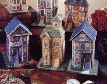 May include: Six miniature houses made of plastic canvas with cross-stitch designs. The houses are in various colors, including purple, blue, green, and red. They are arranged on a table with books and other decorative items.