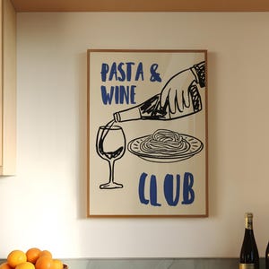 Op de afbeelding: Een keuken met een ingelijste poster met de tekst "Pasta & Wine Club", een kom met sinaasappels en twee wijnflessen. De poster heeft een blauw en wit ontwerp. De sinaasappels zitten in een bruine kom.