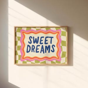 May include: A framed art print with the words "SWEET DREAMS" in blue, set against a cream background with a pink and orange wavy border. The background has a green and white checkered pattern.