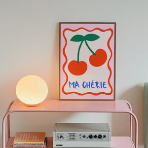 May include: A pink metal shelf with two shelves, a white globe lamp, a stack of books, and a white stereo system. A framed print with a pink background and a graphic of two red cherries with green stems and leaves. The print has the text "MA CH&Eacute;RIE" in blue and red letters.