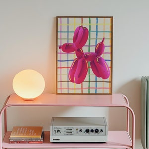 May include: A pink metal side table with two shelves holds a glowing orb lamp, books, and a silver audio device. A framed print of a pink balloon dog against a colorful plaid background hangs on the wall. A green radiator is visible.