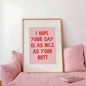 May include: A rectangular framed wall art print featuring a light pink background with bold red hand-lettered text that reads: I HOPE YOUR DAY IS AS NICE AS YOUR BUTT. The print is encased in a simple light-colored wooden frame with a white mat border.