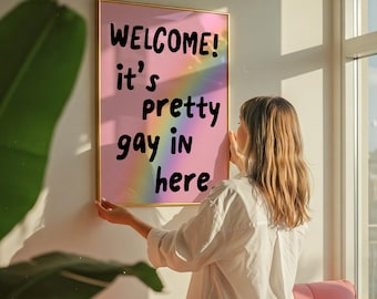 May include: A framed poster with the text "WELCOME! it's pretty gay in here" on a pink background with a rainbow. The poster is being held up against a white wall. The room has a pink chair and a large window.