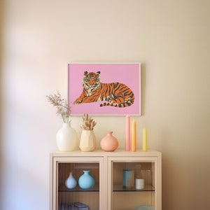 May include: A framed print of an orange and black tiger lying down on a pink background. The print is hanging on a light beige wall above a white shelf with vases and candles.