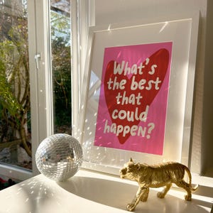 May include: A framed print with the text "What's the best that could happen?" on a pink background with a red heart. A silver disco ball and a gold tiger figurine are on a white surface.