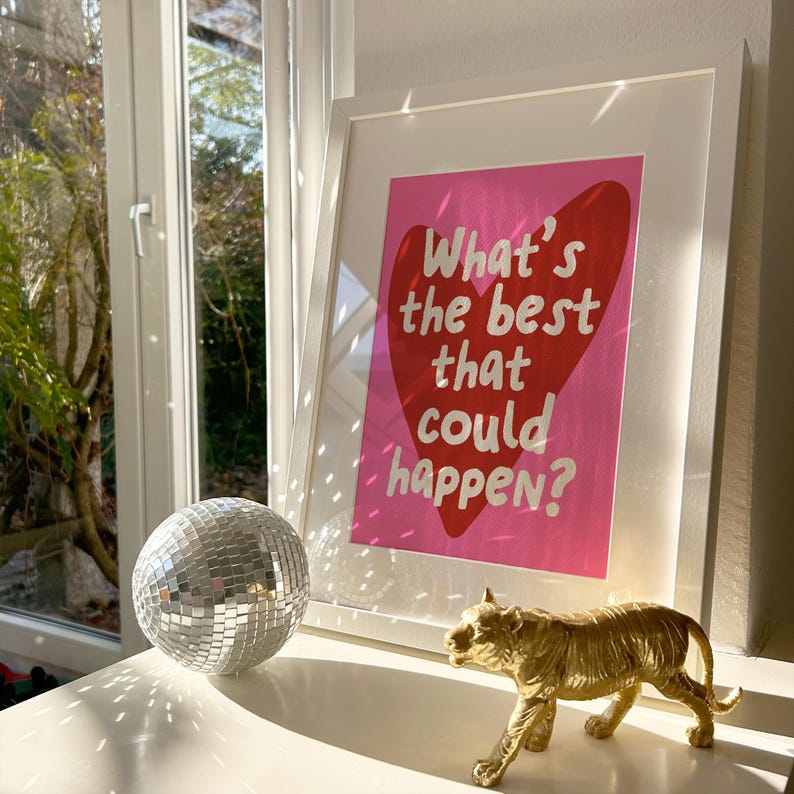 May include: A framed print with the text "What's the best that could happen?" on a pink background with a red heart. A silver disco ball and a gold tiger figurine are on a white surface.