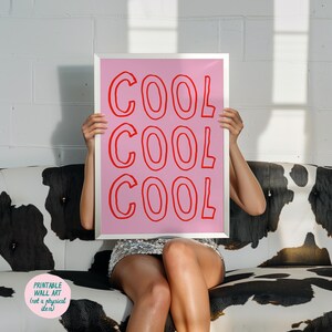 May include: A framed pink print with the word "COOL" repeated three times in red. The print is held up in front of a person sitting on a black and white cow-print couch. The print is labeled "Printable Wall Art (not a physical item)".