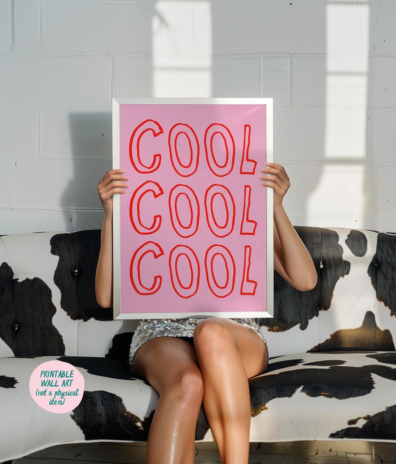 May include: A framed pink print with the word "COOL" repeated three times in red. The print is held up in front of a person sitting on a black and white cow-print couch. The print is labeled "Printable Wall Art (not a physical item)".