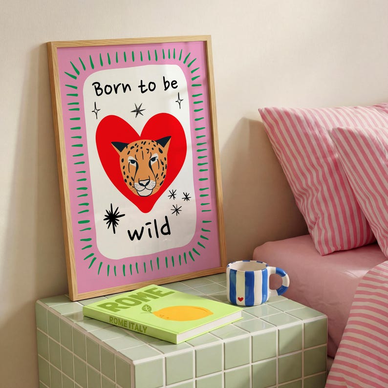 May include: A framed rectangular wall art print featuring a pink border with green dashes, a large red heart, a leopard face illustration, and the black text Born to be wild with star accents. The print is displayed on a tiled surface.