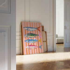 May include: A framed art print displaying four vibrant fish illustrations. The fish are blue, green, blue, and red, with orange and pink details. The background is a pink canvas with vertical orange stripes. The artwork is propped against a wall.