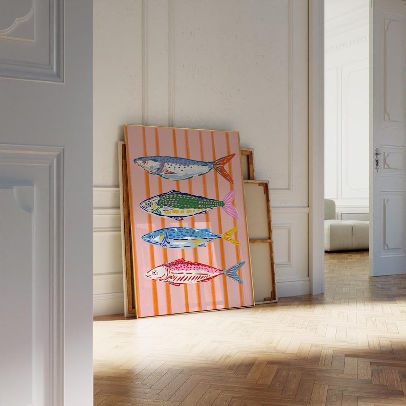 May include: A framed art print displaying four vibrant fish illustrations. The fish are blue, green, blue, and red, with orange and pink details. The background is a pink canvas with vertical orange stripes. The artwork is propped against a wall.