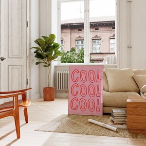 May include: A pink framed print with the word "COOL" repeated three times in red. The print leans against a beige sofa in a room with a neutral colour scheme. A potted plant and wooden chair are in the foreground.