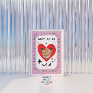 May include: A framed wall art print featuring a white background with a large red heart in the centre containing a leopard face. The text Born to be wild is written in black above and below the heart, surrounded by a pink border with green dashes.