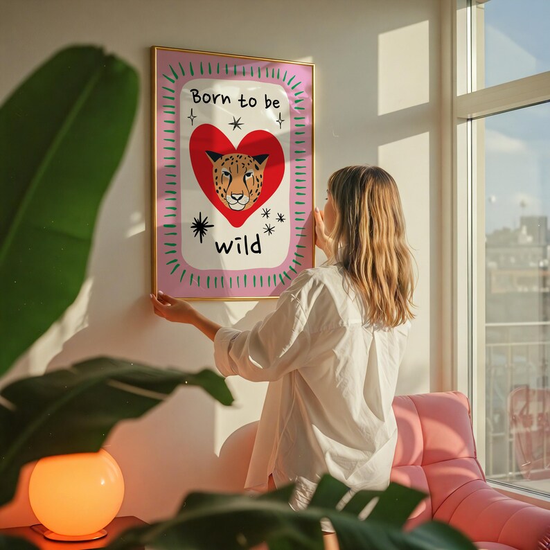 May include: A rectangular wall art print in a gold frame featuring a red heart with a leopard face inside. The text Born to be wild is printed in black. The background is white with a pink border and green dashes, accented by black star icons.