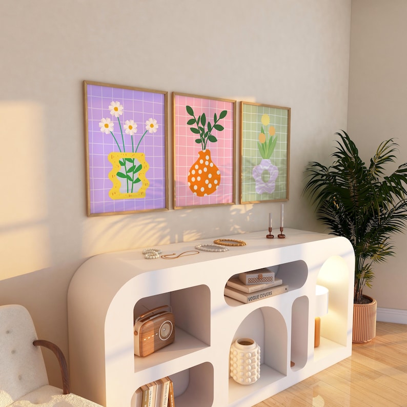 May include: Three framed art prints with floral designs. Each print features a vase with flowers or foliage against a grid background. The color palette includes purple, pink, and green, with wooden frames. The prints are displayed above a white cabinet.