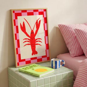 Puede incluir: Un cuadro enmarcado con una ilustración de langosta roja sobre un fondo blanquecino, enmarcado por un borde a cuadros rosa y rojo. Una taza a rayas azules y blancas con un corazón está al lado de un libro titulado "ROME ITALY".