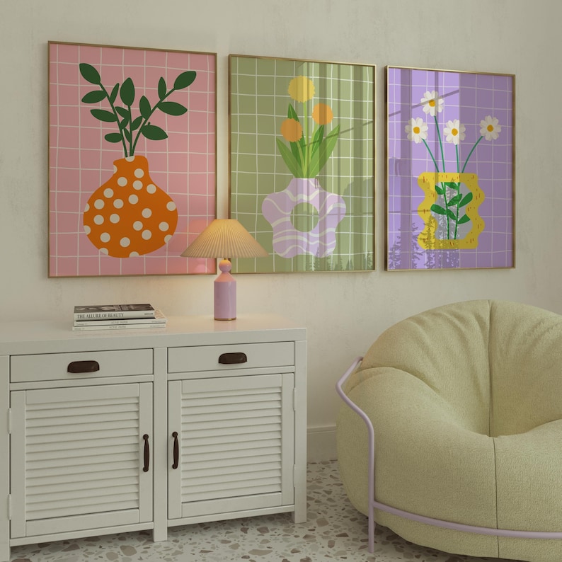 May include: Three framed art prints with floral designs. The prints depict vases with flowers and foliage against a grid background. The colour palette includes pink, green, and purple, with gold frames. A lamp and a white cabinet are in the foreground.
